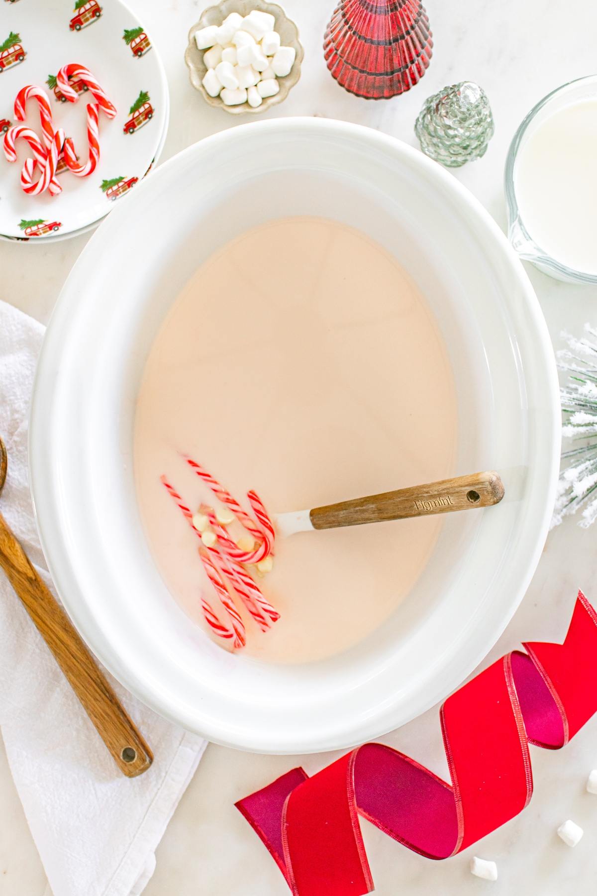 White slow cooker with creamy liquid, candy canes, and spoon, surrounded by Christmas decor and treats.