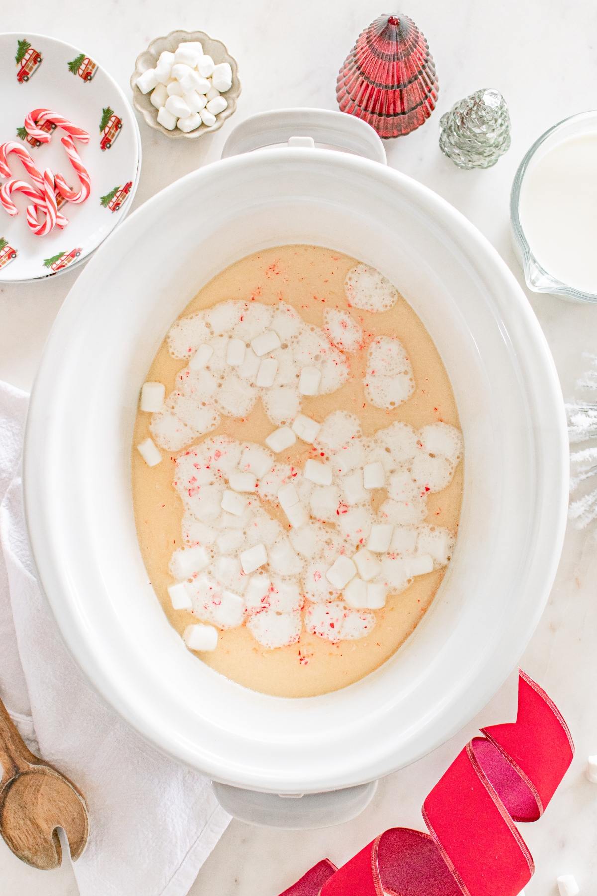 A slow cooker with white hot chocolate, topped with marshmallows, surrounded by holiday decorations.