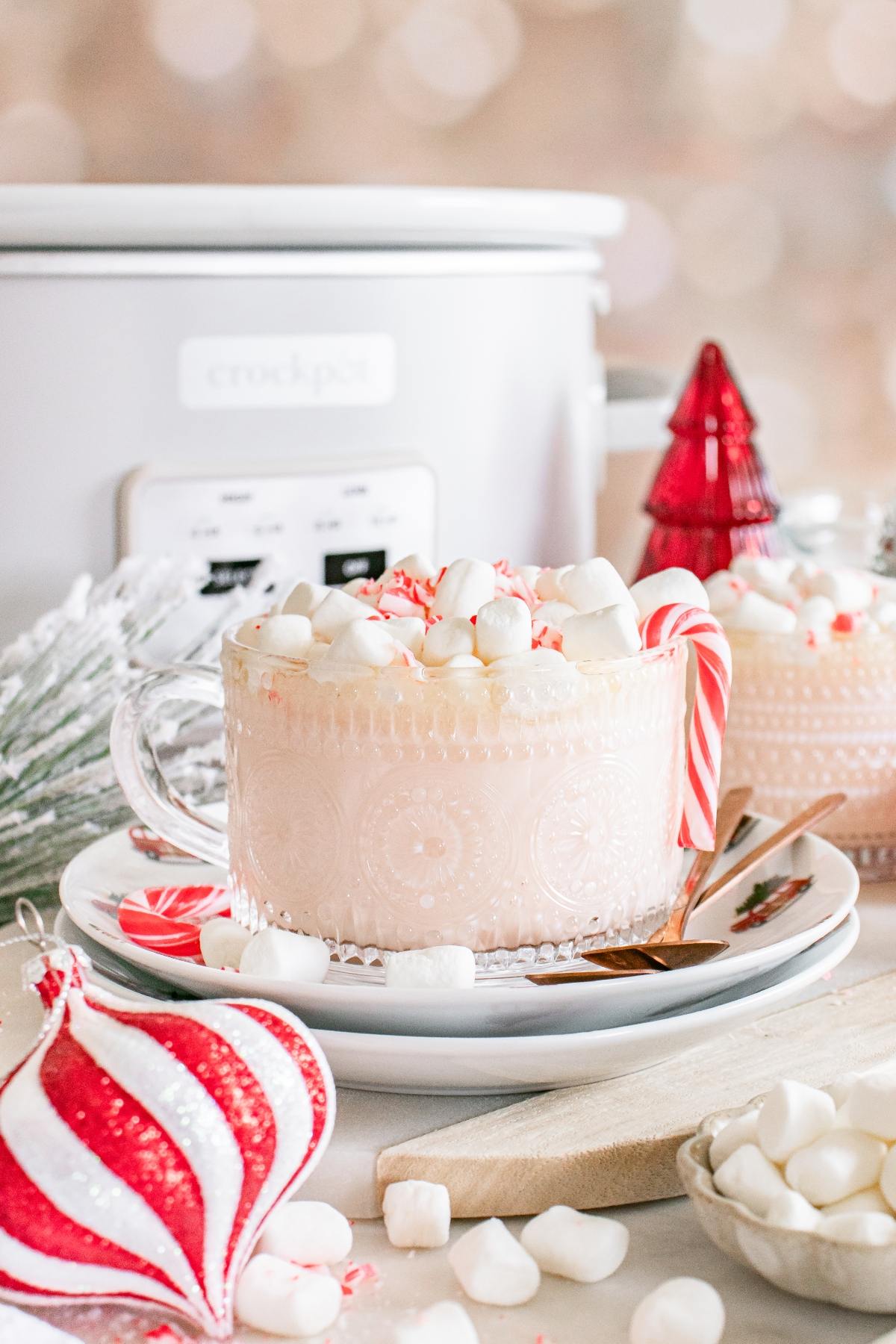 A mug of white hot chocolate with marshmallows and a candy cane, surrounded by festive holiday decorations.