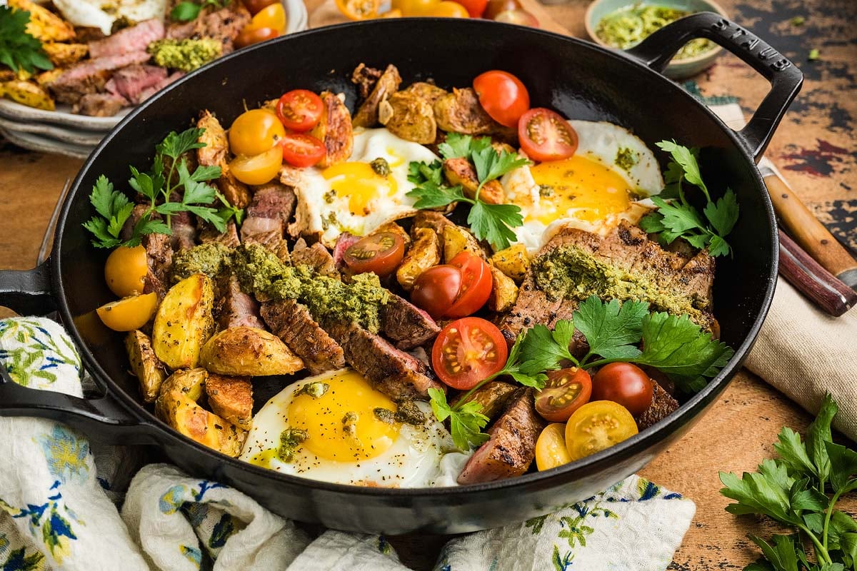 A hearty skillet featuring steak and eggs, golden roasted potatoes, juicy tomatoes, fresh herbs, and a vibrant green sauce.