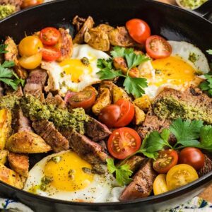 A skillet with steak and eggs, roasted potatoes, tomatoes, herbs, and green sauce.