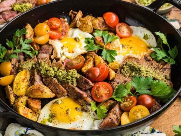A skillet with steak and eggs, roasted potatoes, tomatoes, herbs, and green sauce.