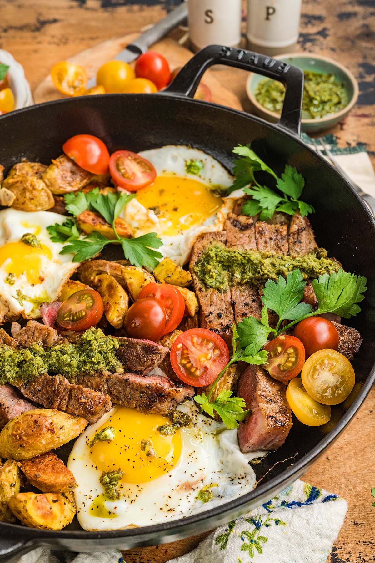 A hearty skillet with steak and eggs, roasted potatoes, cherry tomatoes, fresh herbs, and a vibrant green sauce.