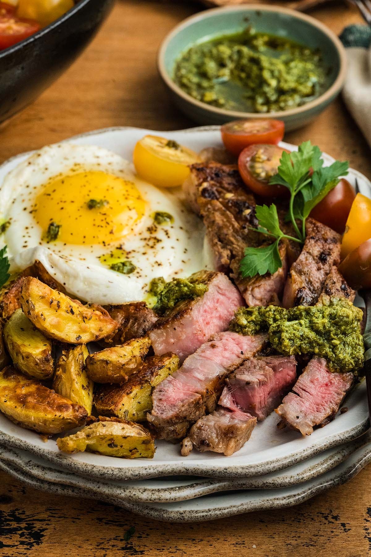 Plate with steak and eggs, fried potatoes, sunny-side up egg, cherry tomatoes, and green sauce garnish.