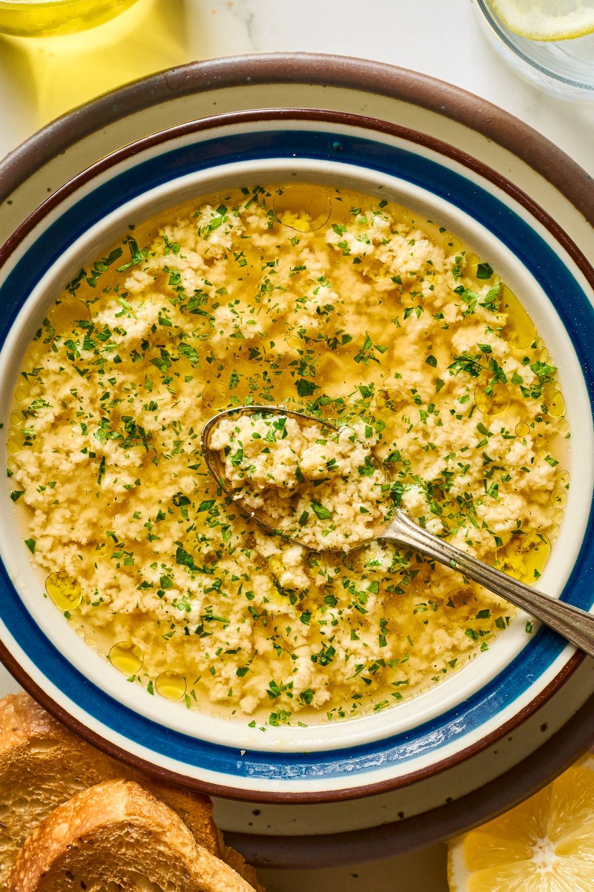 A bowl of egg drop stracciatella soup garnished with herbs, with a spoon and toasted bread on the side.