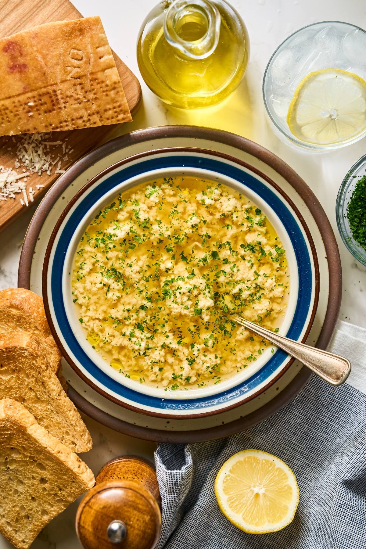 A bowl of egg drop stracciatella soup with herbs, surrounded by bread, cheese, lemon, and a bottle of olive oil.