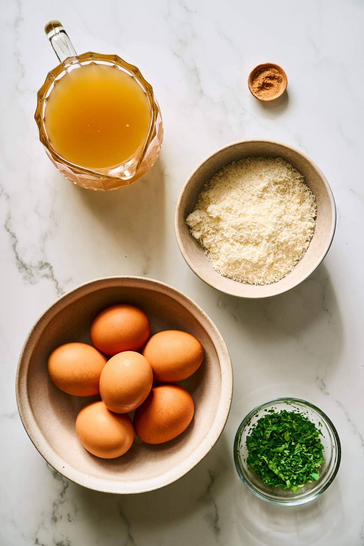 Eggs, grated cheese, chopped herbs, broth, and a small bowl of spice on a marble surface.