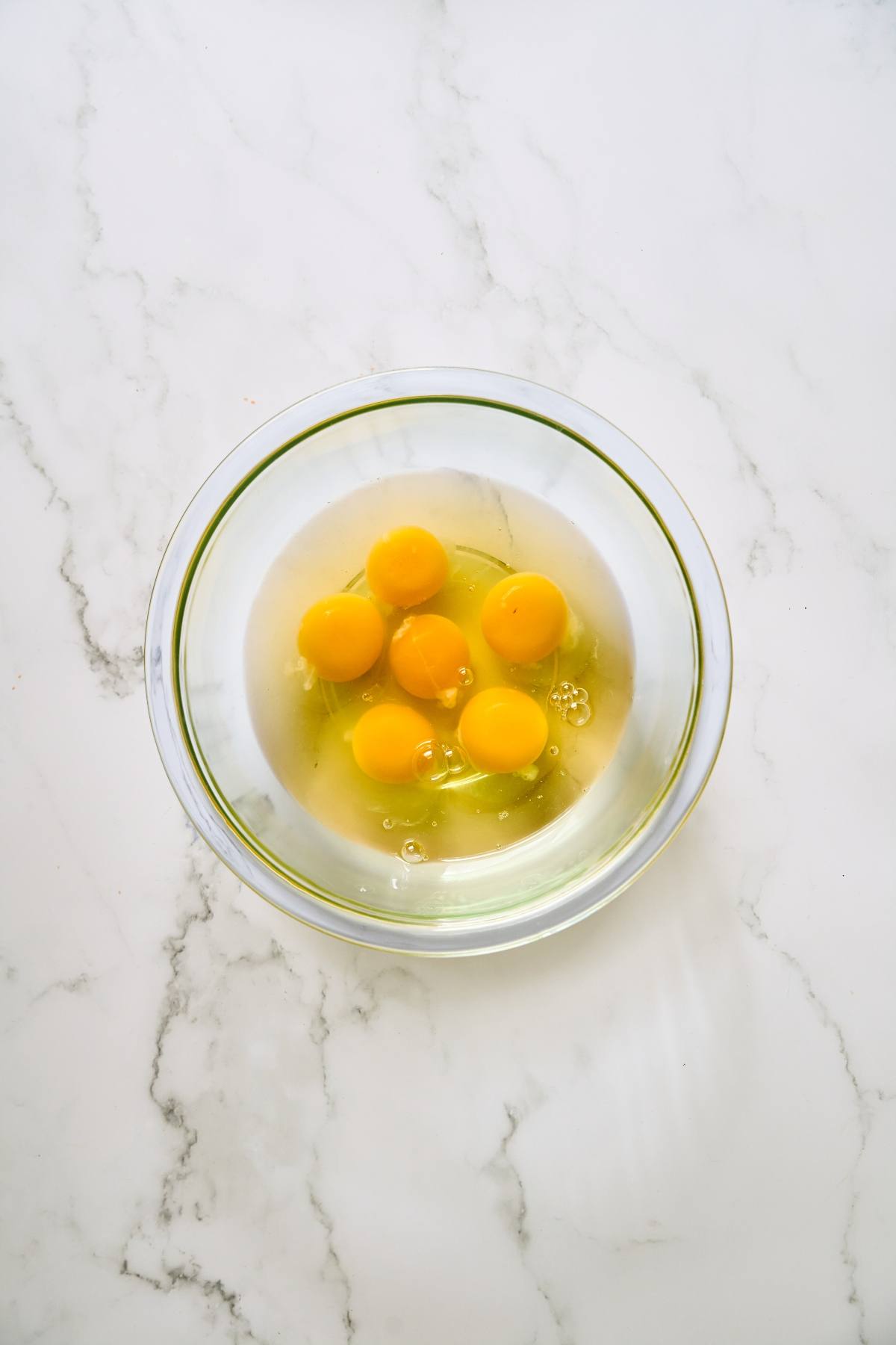 A glass bowl with six raw eggs cracked open on a white marble surface.