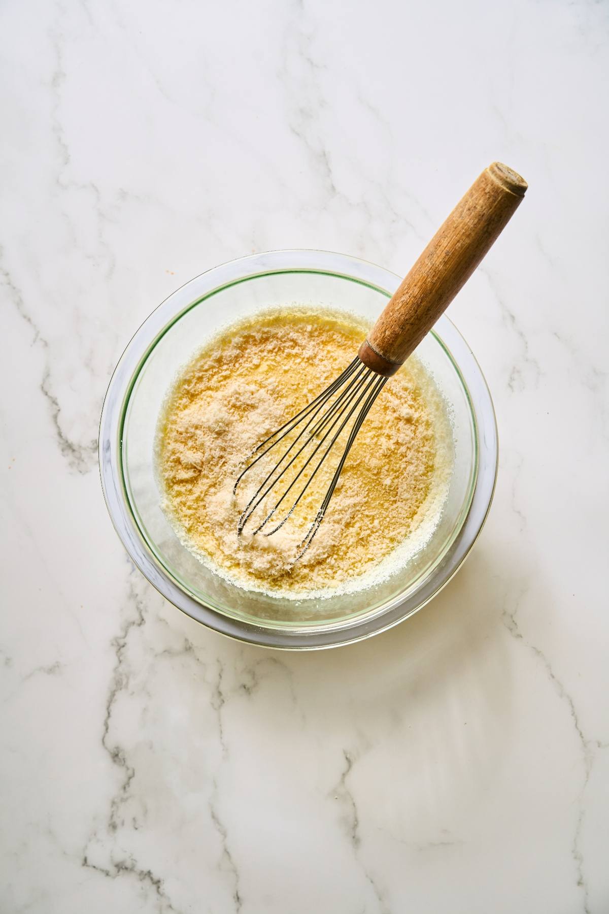 A whisk in a glass bowl mixing dry ingredients on a white marble surface.