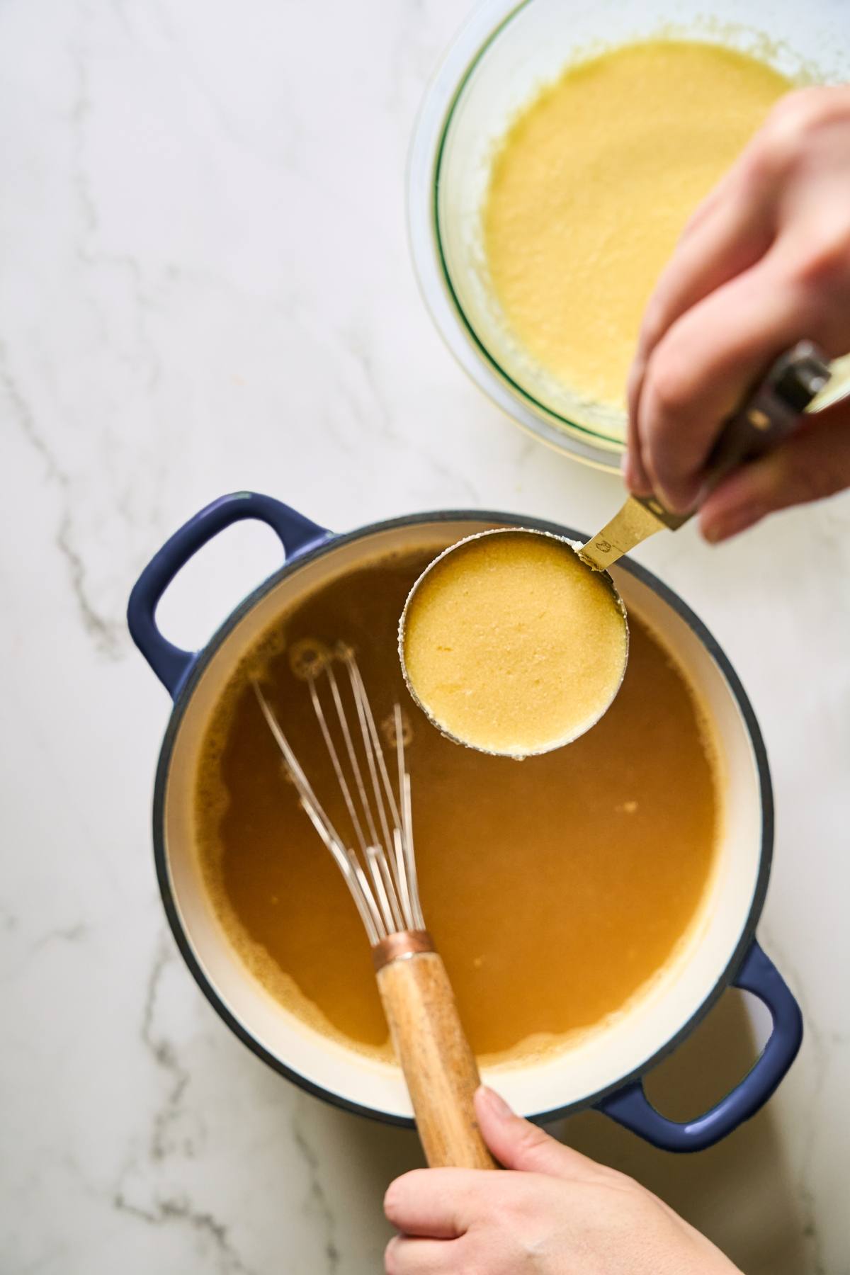 A hand pours yellow cornmeal into a pot of liquid while whisking, with a bowl of cornmeal nearby.