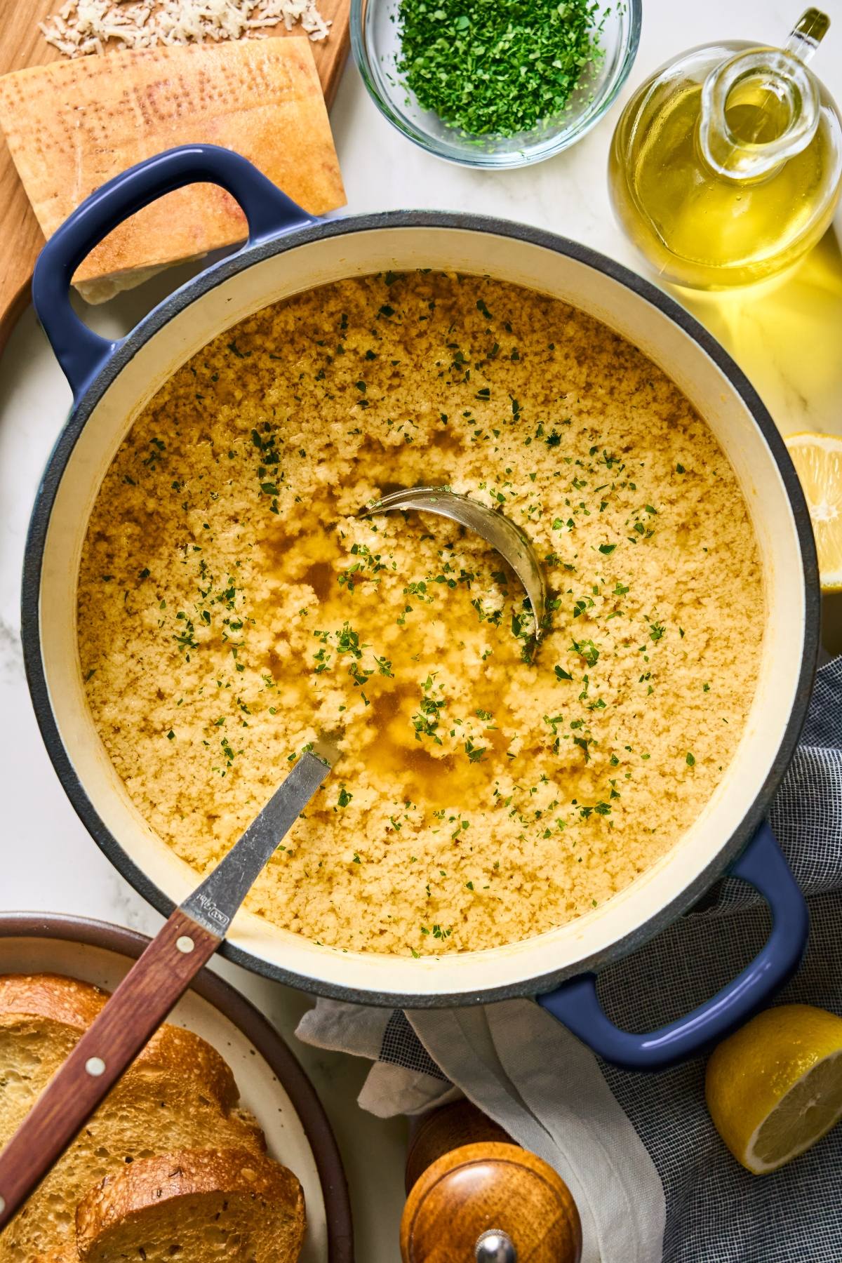 A pot of stracciatella soup garnished with herbs, with a ladle inside, surrounded by bread and cooking ingredients.