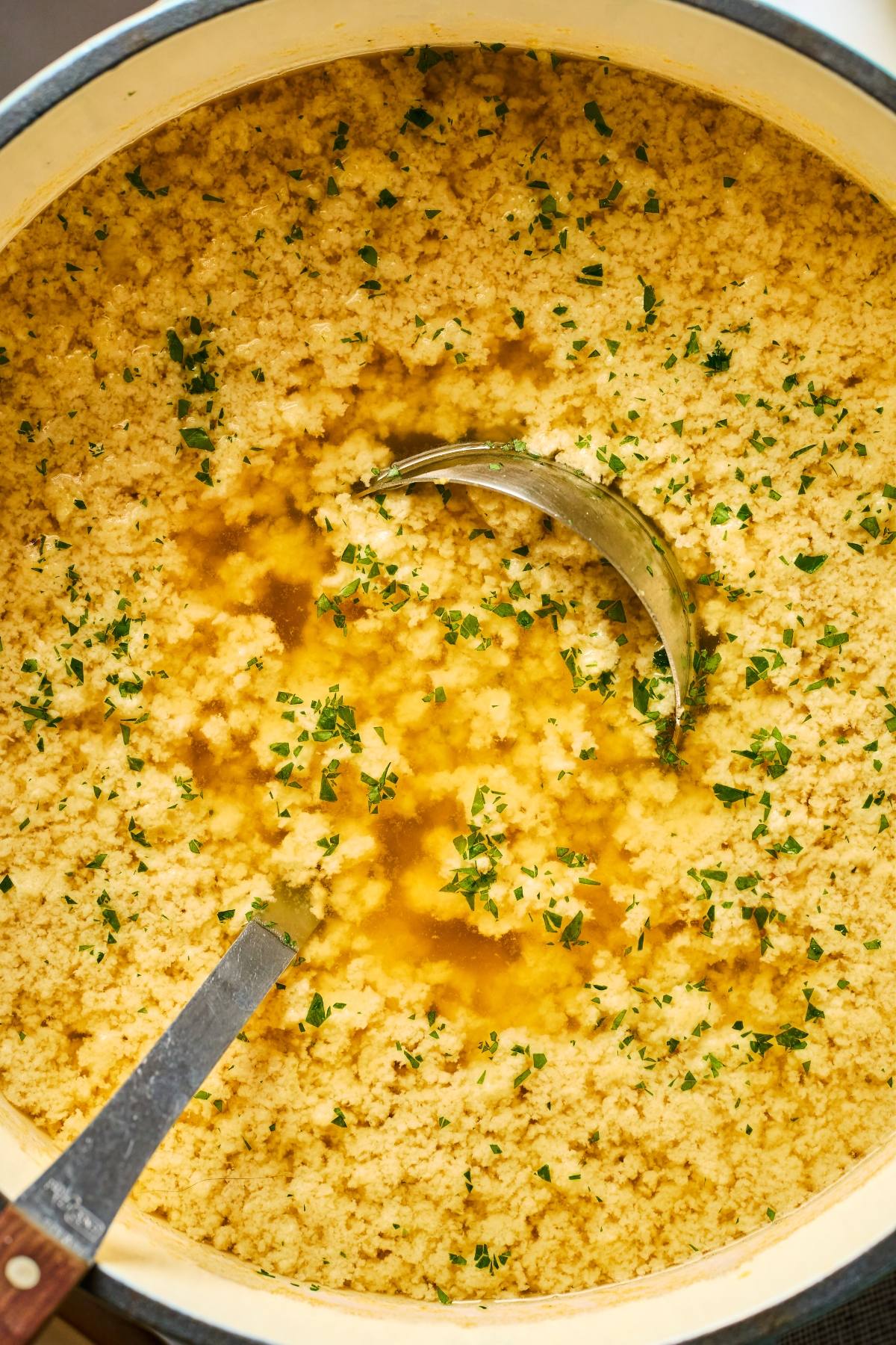 A pot of couscous with broth, herbs, and a ladle partially submerged in the mixture.