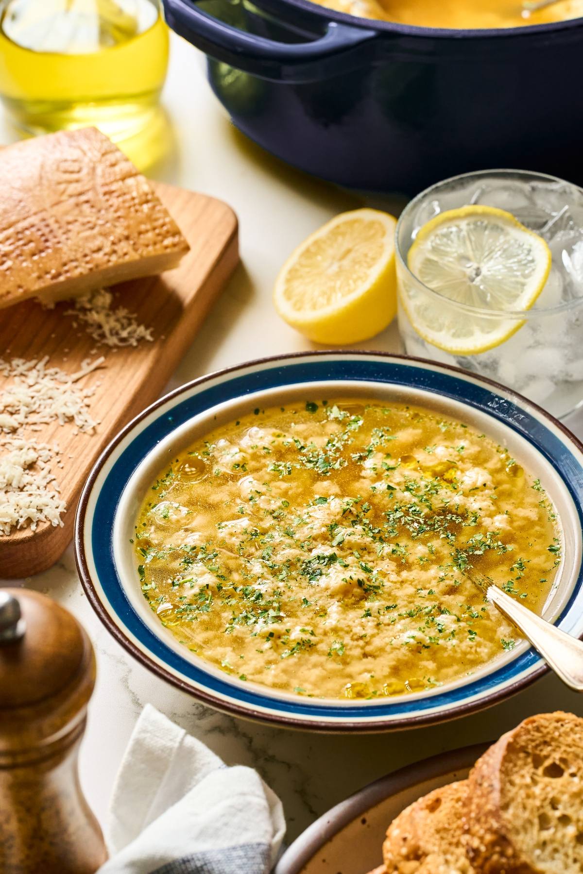 A bowl of stracciatella soup garnished with herbs, with bread, lemon, and a glass of water on the table.