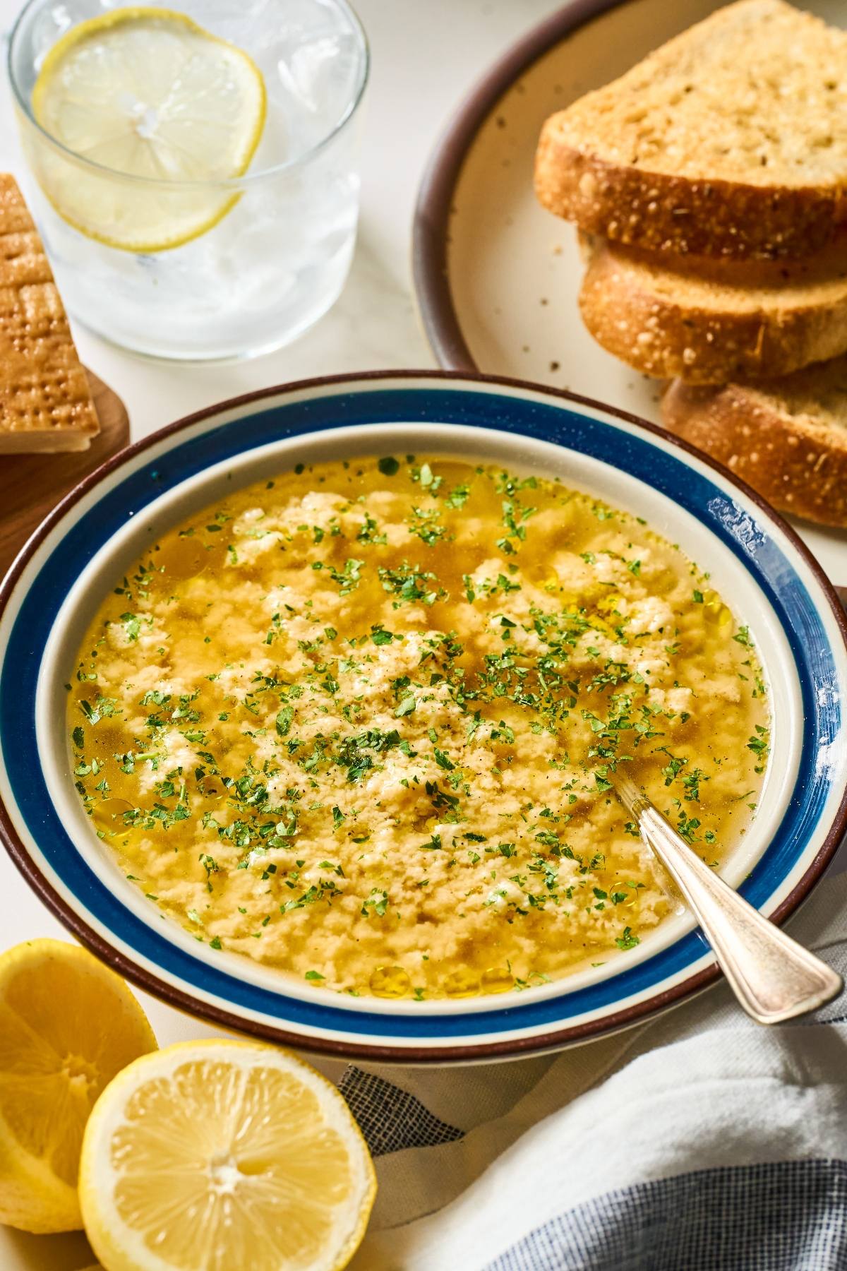 A bowl of egg drop stracciatella soup garnished with herbs, served with lemon and slices of toasted bread.