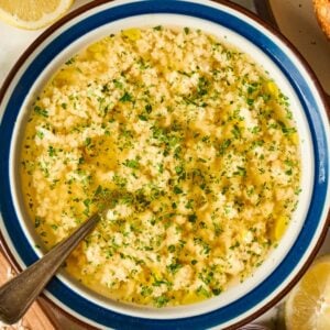 A bowl of garlic spread topped with herbs and olive oil, surrounded by bread, lemon, and grated cheese—perfect as a starter alongside Stracciatella Soup.