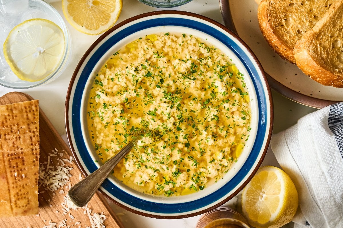 Bowl of Stracciatella Soup garnished with herbs, surrounded by bread, lemon slices, and cheese.