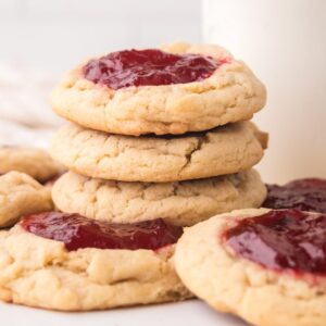 A stack of Strawberry Thumbprint Cookies with red jam centers sits next to a glass of milk on a white surface.