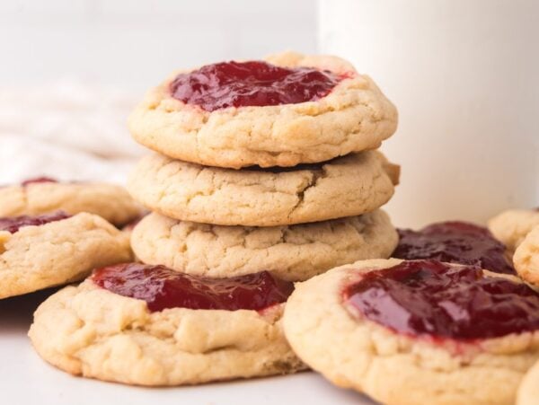 A stack of Strawberry Thumbprint Cookies with red jam centers sits next to a glass of milk on a white surface.