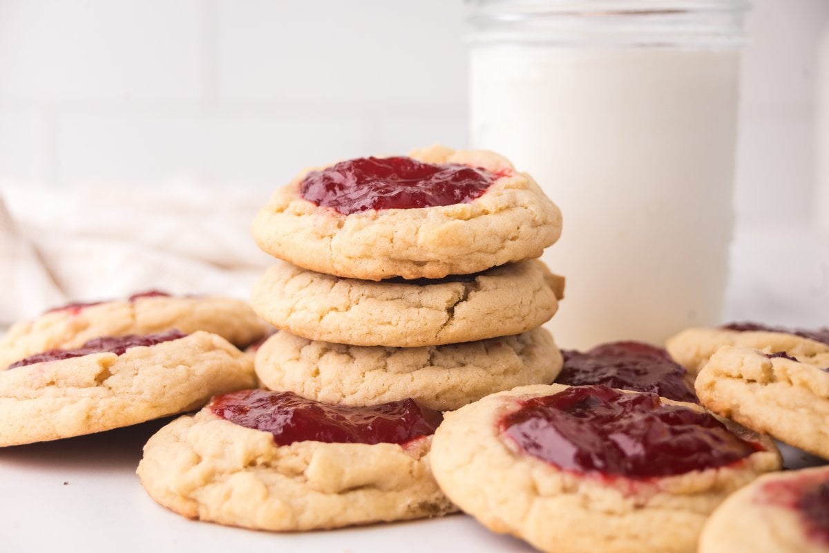 A stack of Strawberry Thumbprint Cookies with red jam centers sits next to a glass of milk on a white surface.