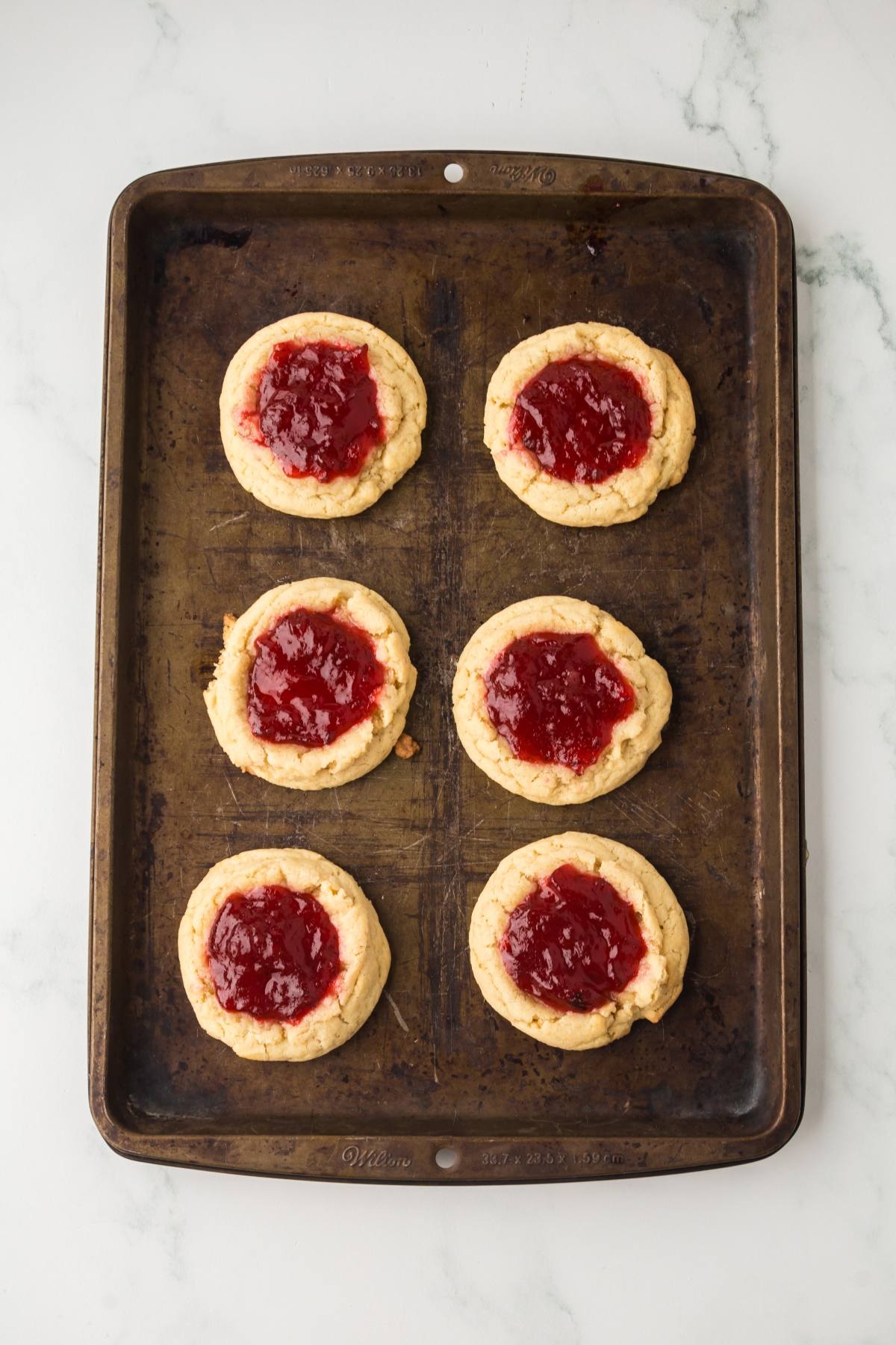 A baking sheet with six jam-filled thumbprint cookies on a white marble surface.