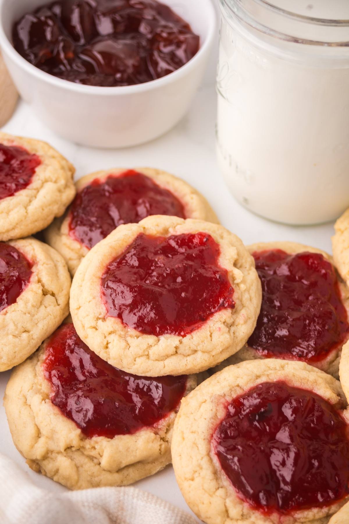 Strawberry Thumbprint cookies topped with red jam filling, next to a jar of milk and a bowl of extra jam.