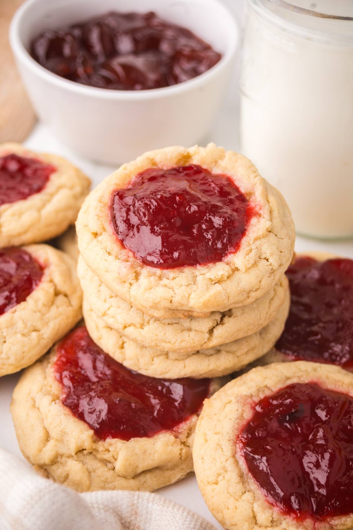 Stack of thumbprint cookies with red jam filling, a glass of milk, and a bowl of jam in the background.