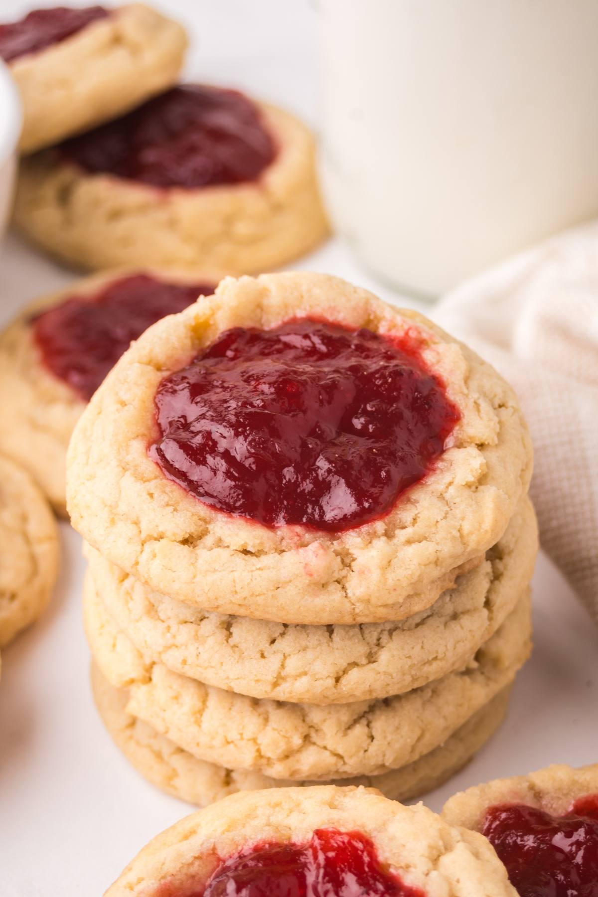 Stack of cookies with red jam centers, with more cookies and a glass of milk in the background.