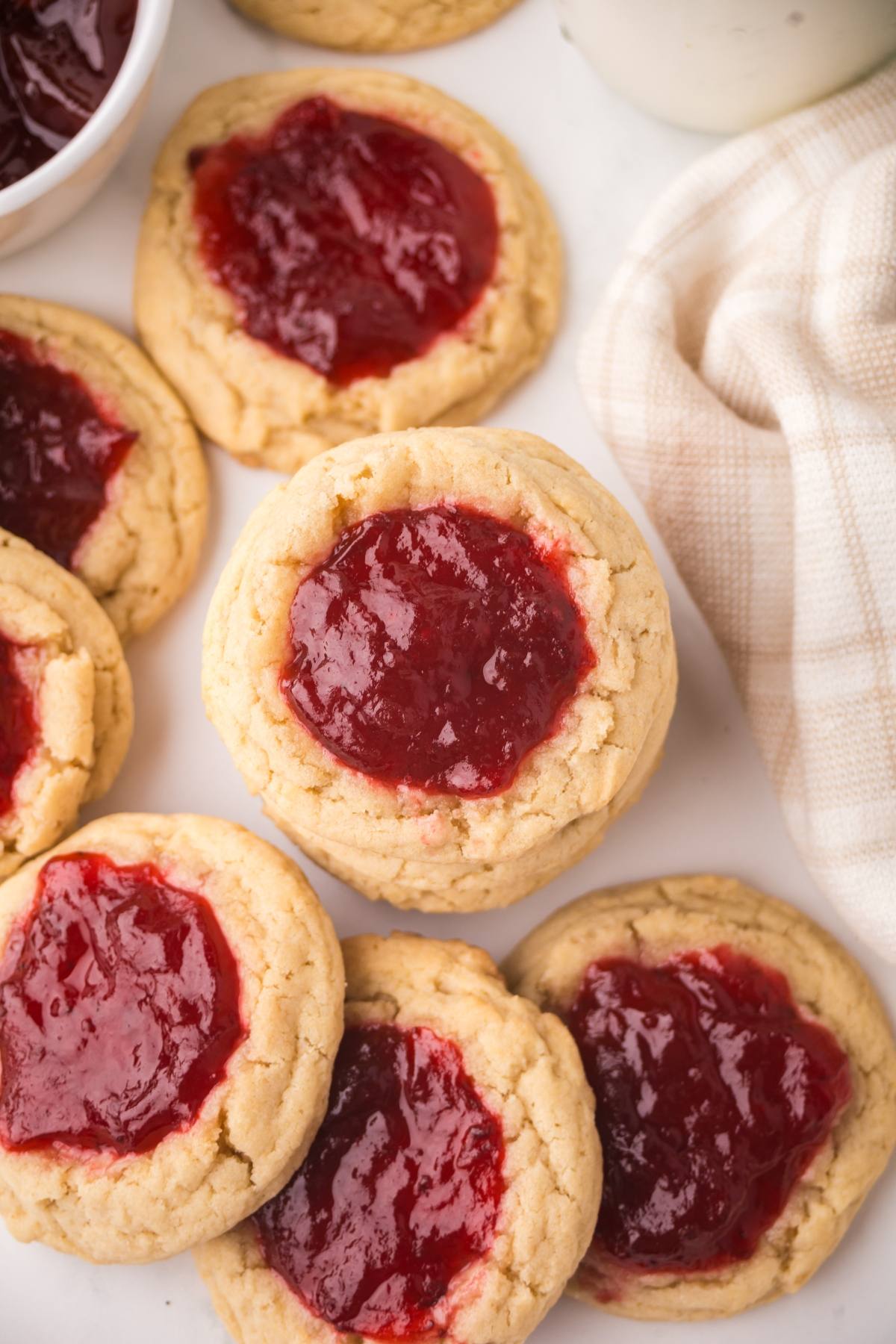 A plate of thumbprint cookies filled with red jam, next to a beige checkered cloth.