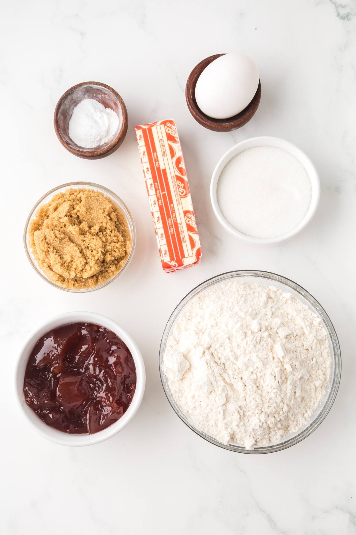 Baking ingredients: flour, butter, egg, salt, sugar, brown sugar, and strawberry jam on a white surface.