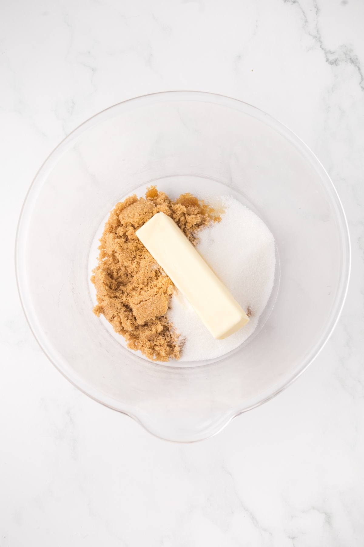 A glass bowl with butter, brown sugar, and white sugar on a white marble surface.