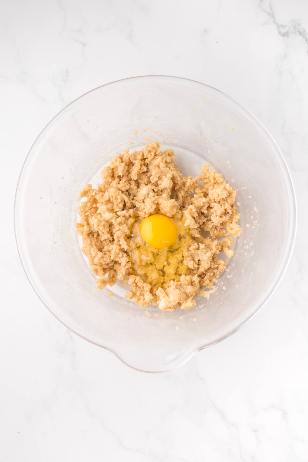 A glass bowl with cookie dough mixture and an egg on top, ready to be mixed, on a white surface.
