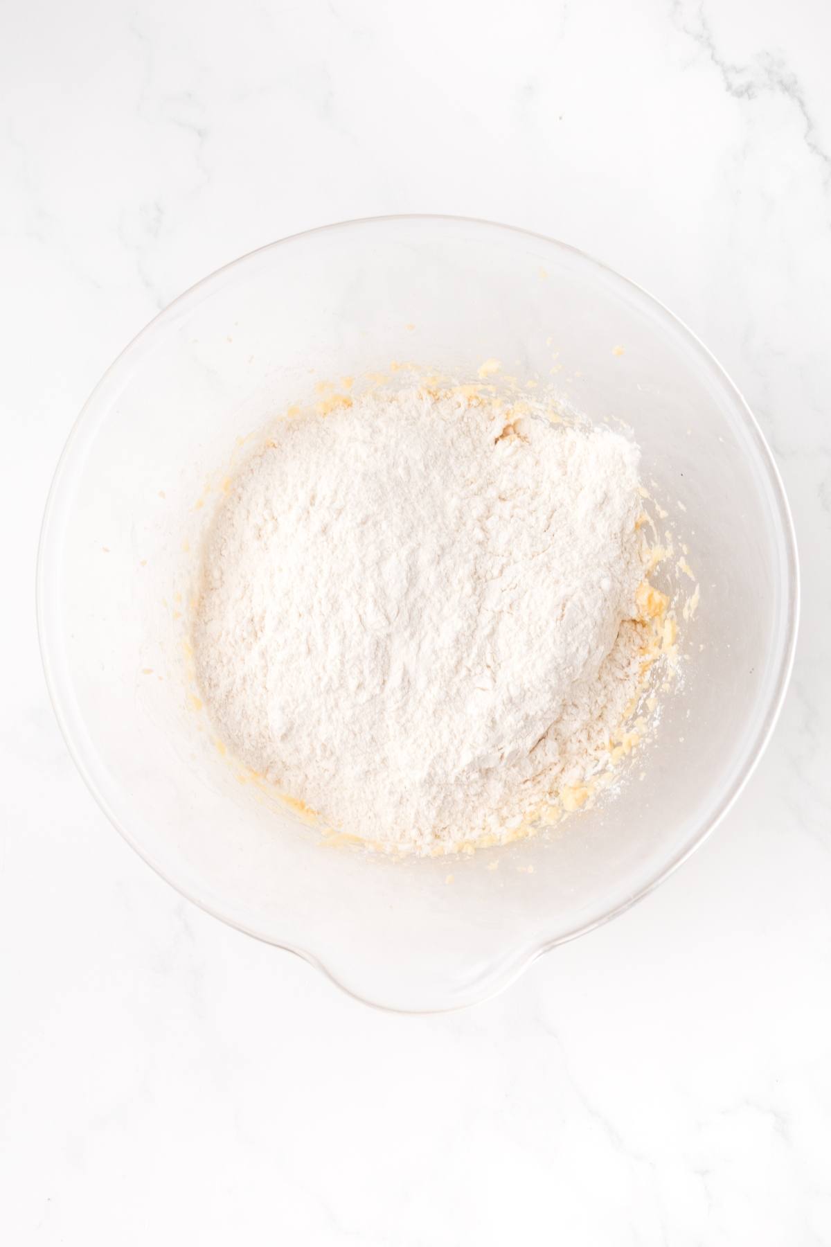 A glass mixing bowl with flour and batter on a white marble surface.
