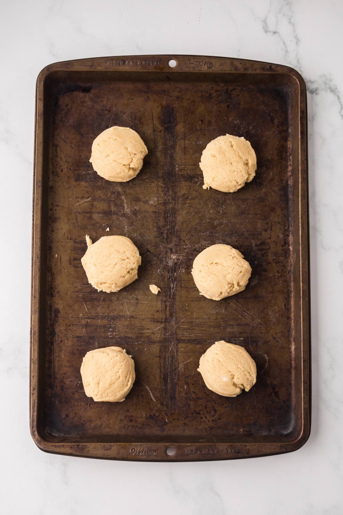 Six balls of cookie dough spaced evenly on a dark baking sheet, ready to be baked.