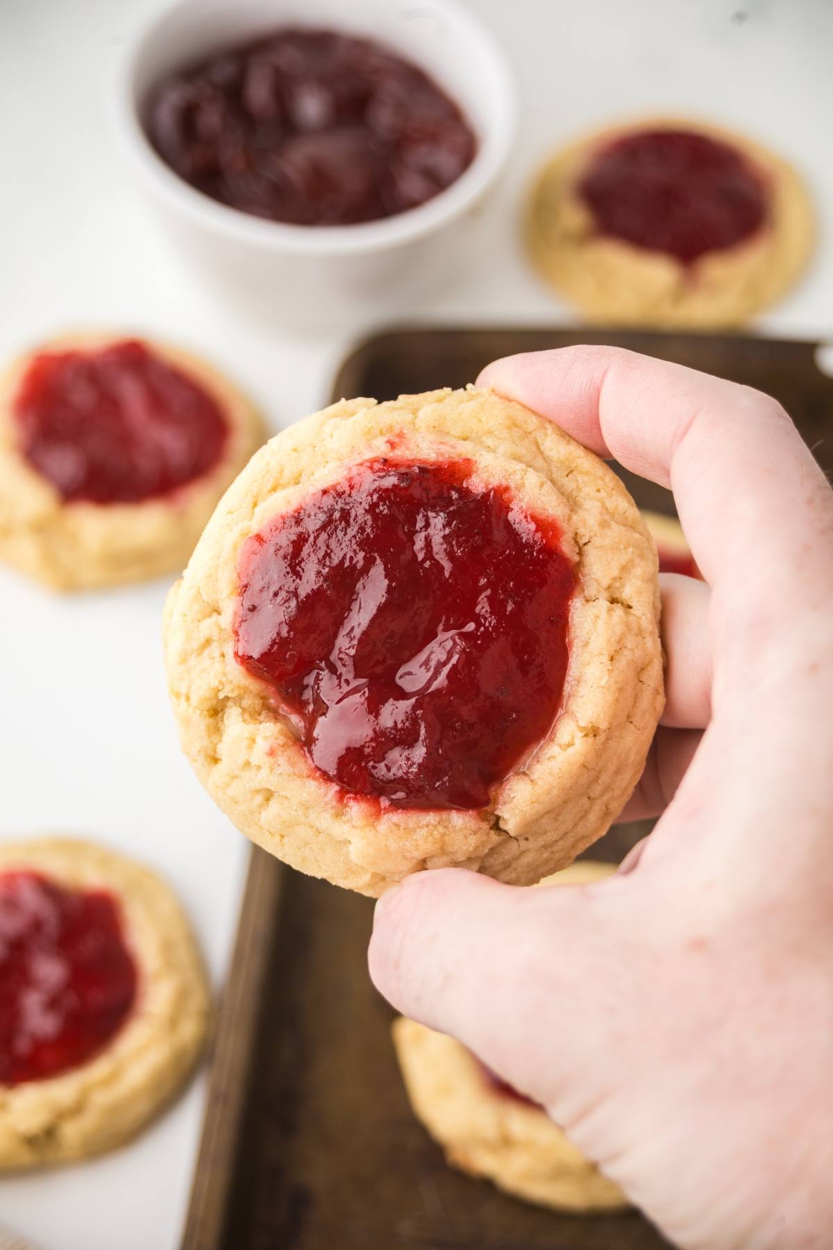 A hand holds a cookie topped with red jam; more jam-topped cookies and a bowl of jam are in the background.