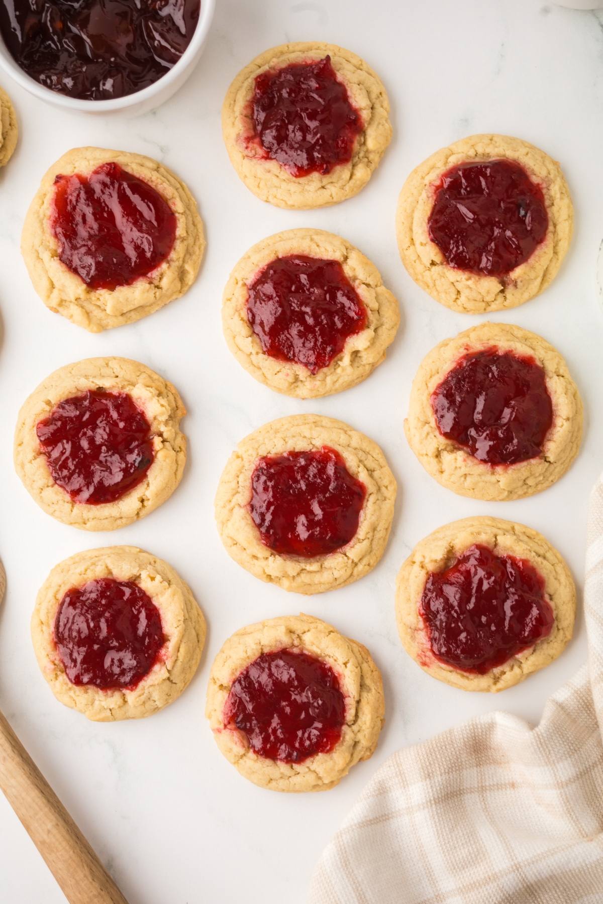 Twelve cookies topped with red jam are arranged on a white surface next to a bowl of jam and a beige cloth.