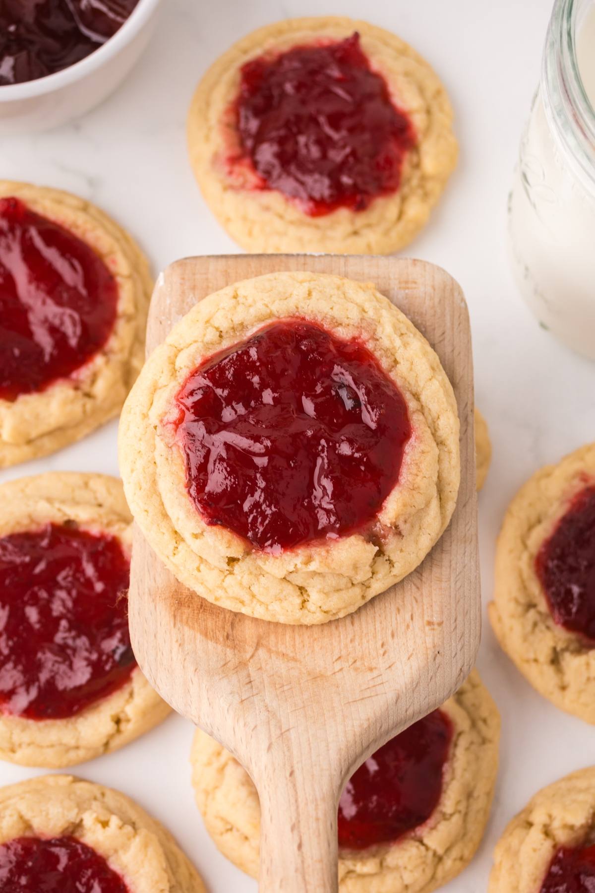 Close-up of cookies topped with red jam, one on a wooden spatula, surrounded by more cookies and a glass of milk.