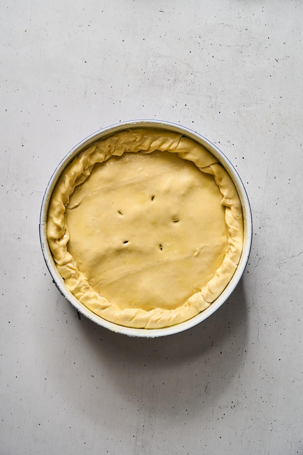 Unbaked pie with a crimped edge and three slits in the top crust, in a round pan on a light surface.