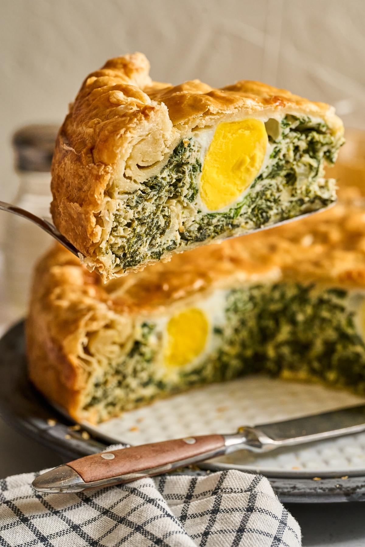 A slice of Torta Pasqualina pie with spinach, egg, and flaky crust being lifted from a pie dish.