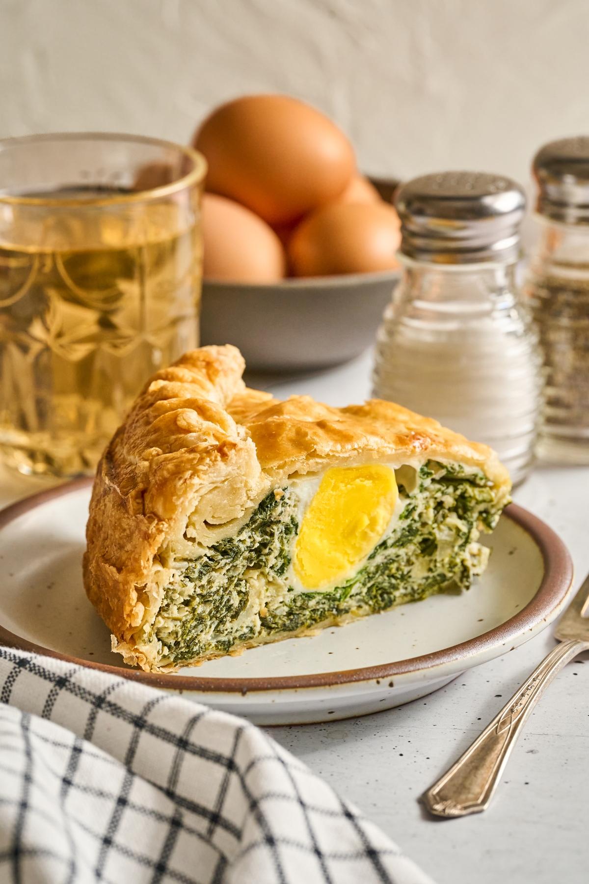 A slice of savory pie with spinach and egg, next to salt, pepper, a glass of drink, and a bowl of eggs.