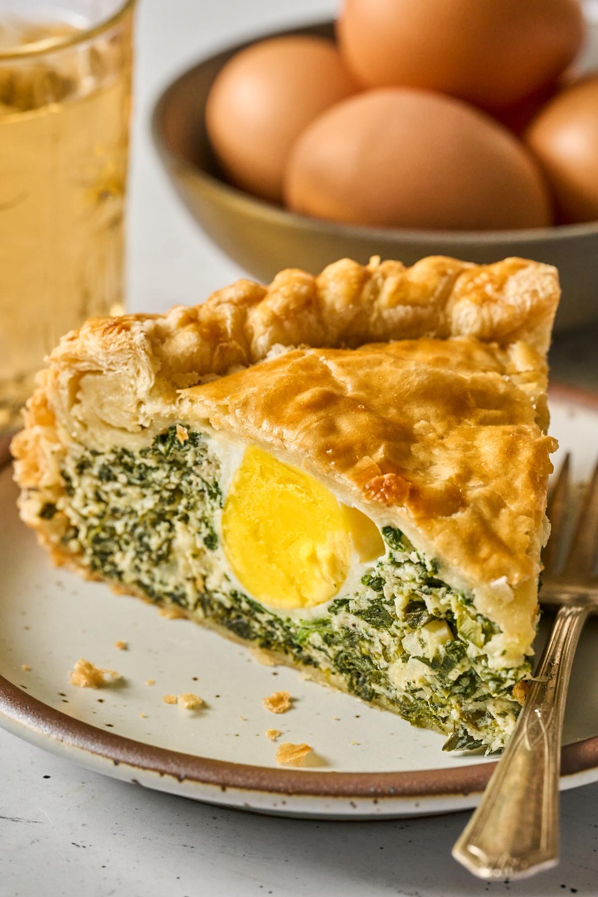 A slice of Torta Pasqualina pie with spinach and a whole egg inside, served on a plate with a fork; eggs in background.