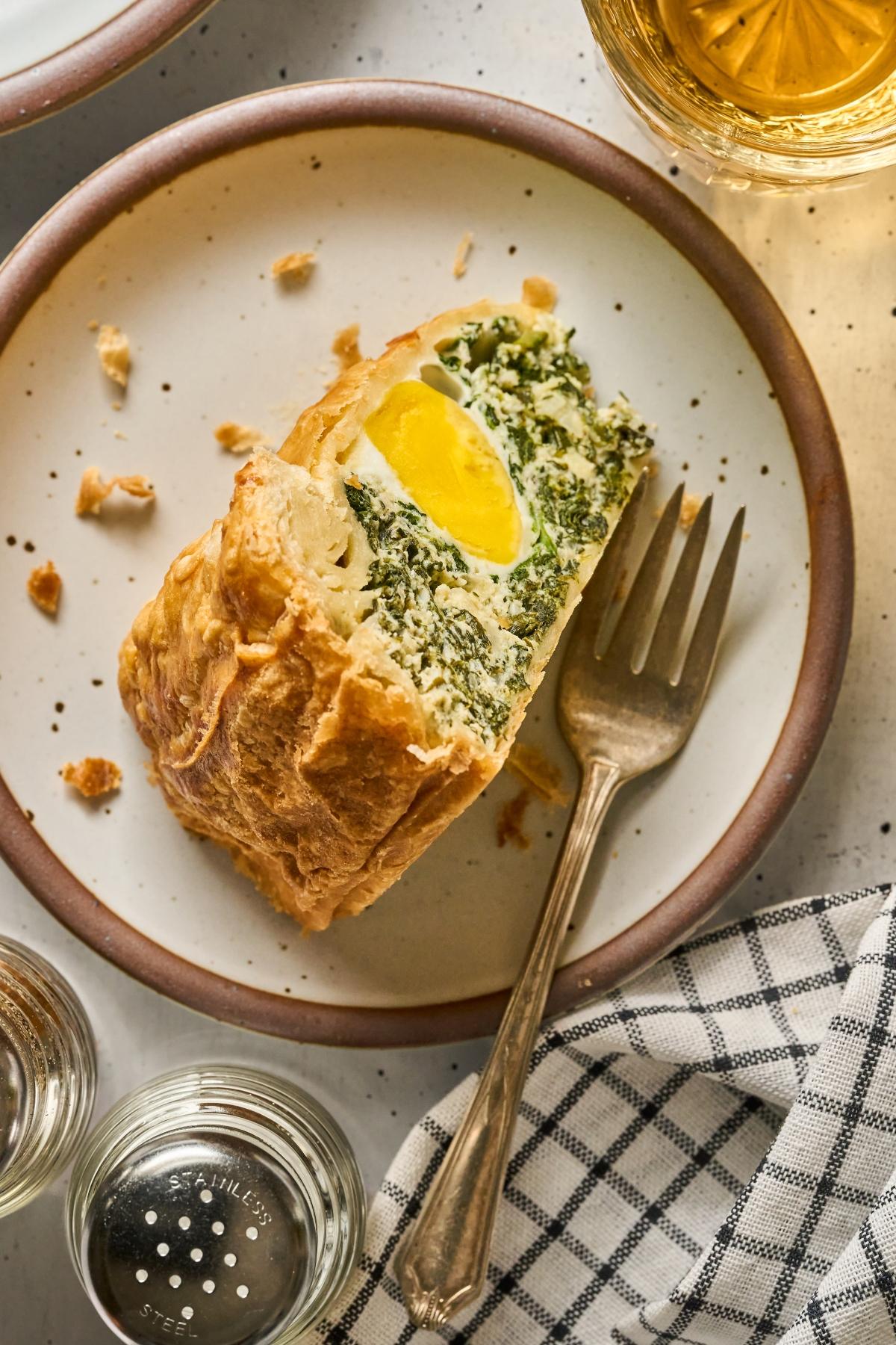A slice of pastry with egg and greens on a plate with a fork, next to a drink and a checkered napkin.