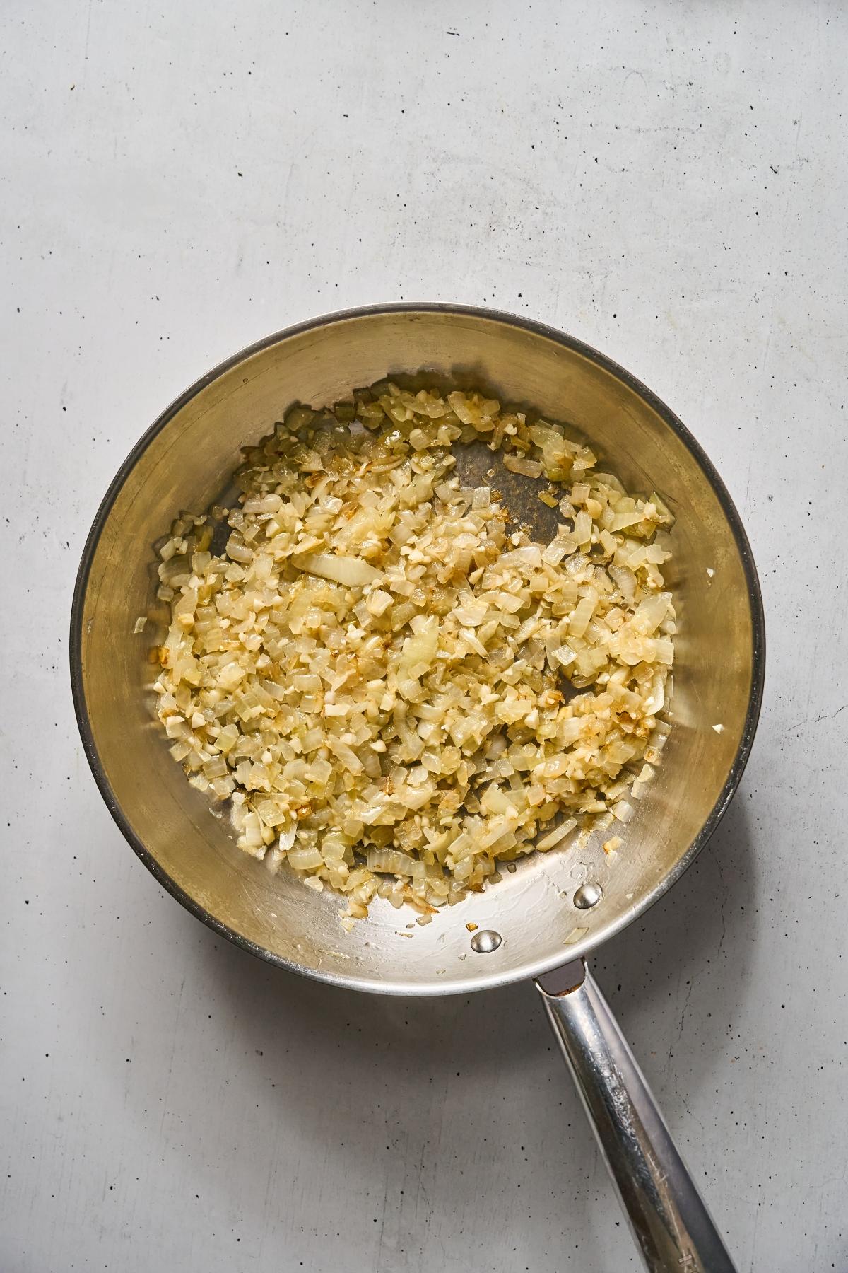 Chopped onions being sautéed in a stainless steel pan on a light-colored surface.