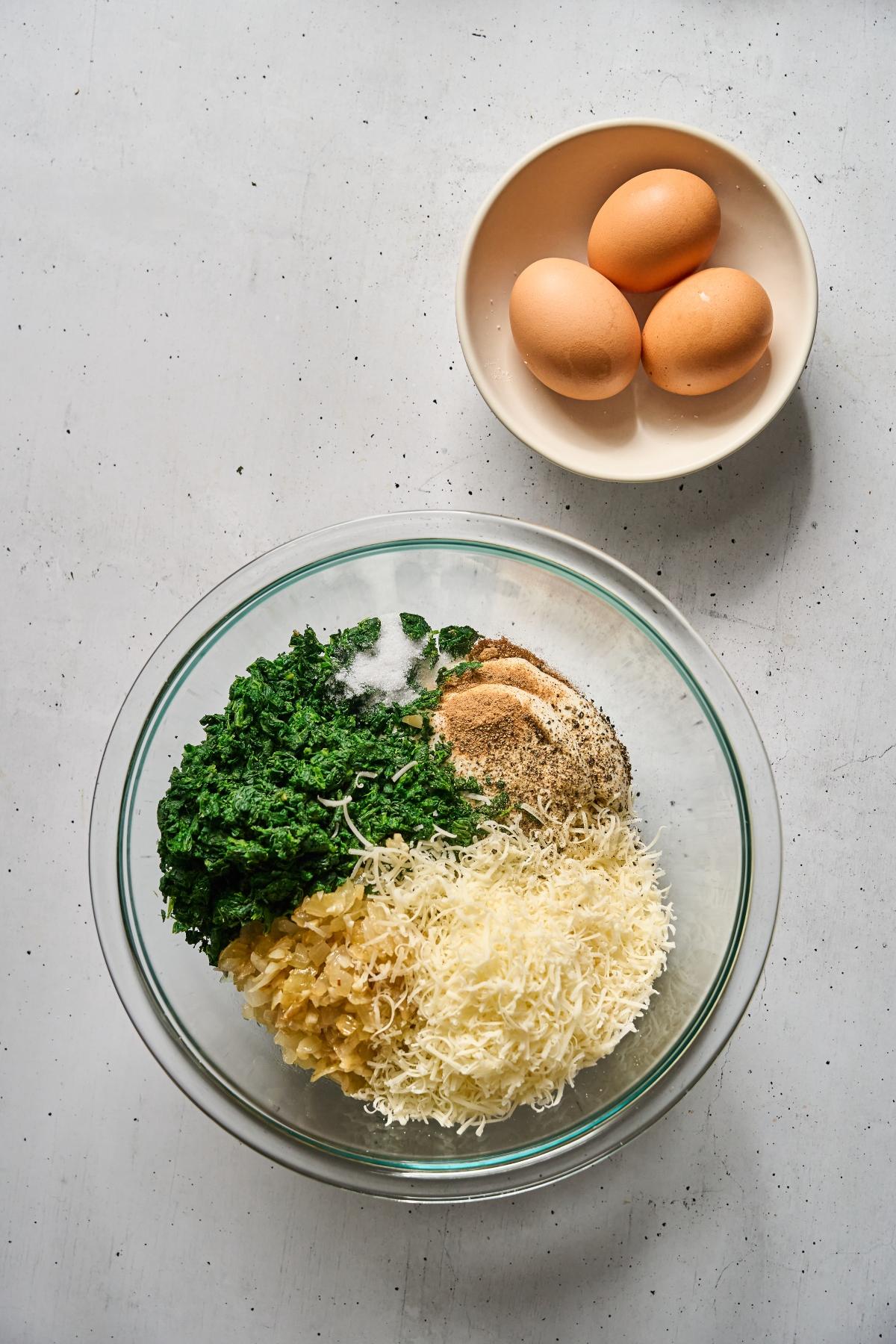 A bowl with chopped spinach, grated cheese, breadcrumbs, and seasonings next to a plate with three eggs.