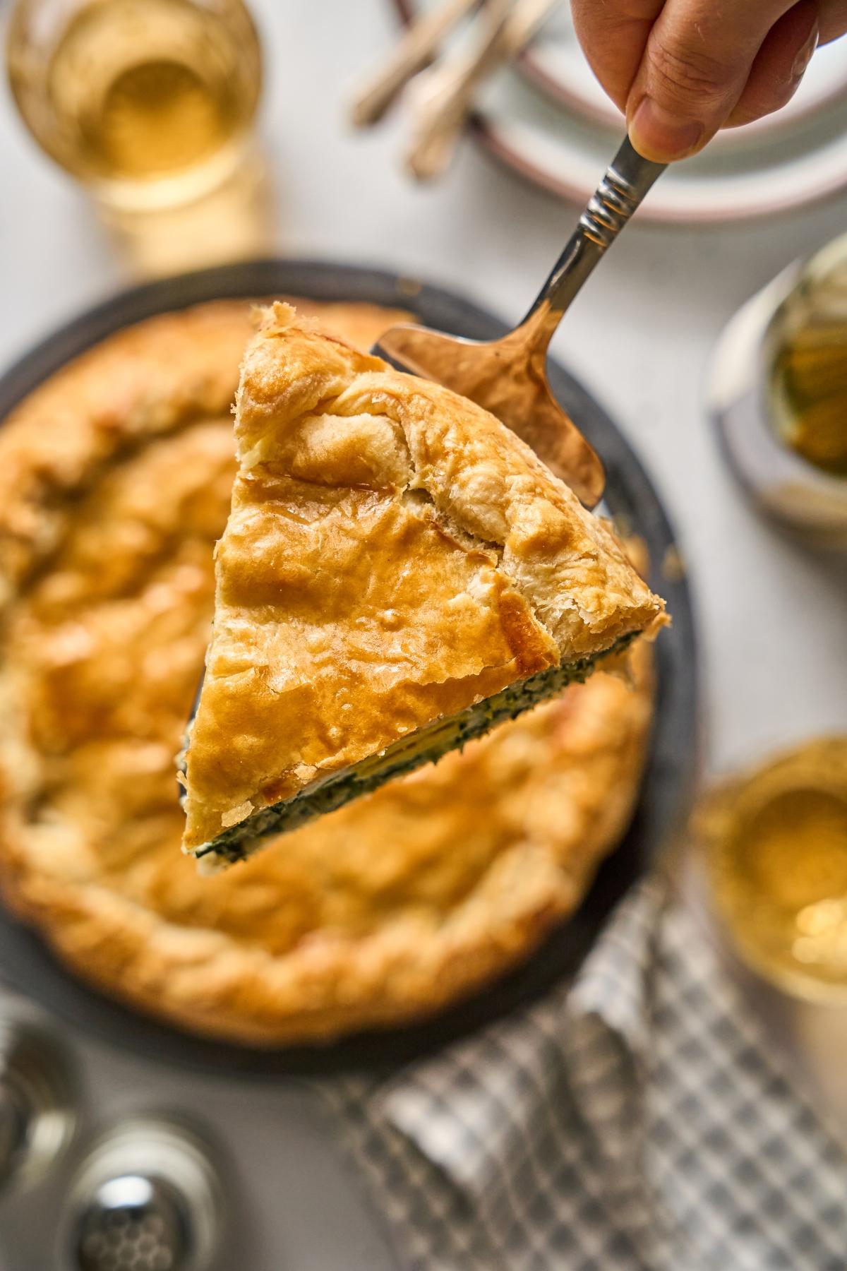 A hand holds up a slice of golden, flaky pie filled with greens above a whole pie on a table.