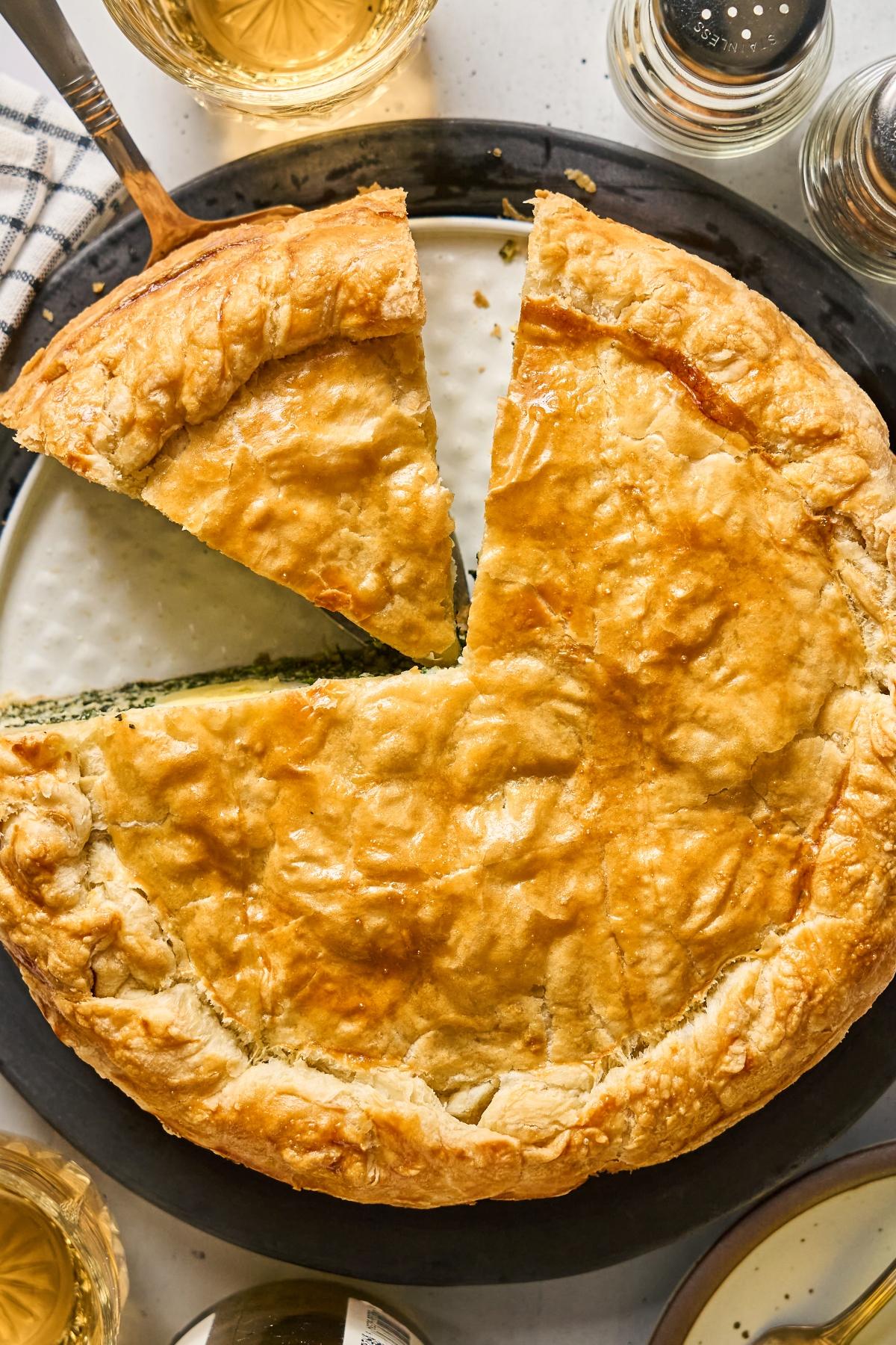 Golden brown savory pie with a slice cut out, surrounded by drinks and a checkered napkin.