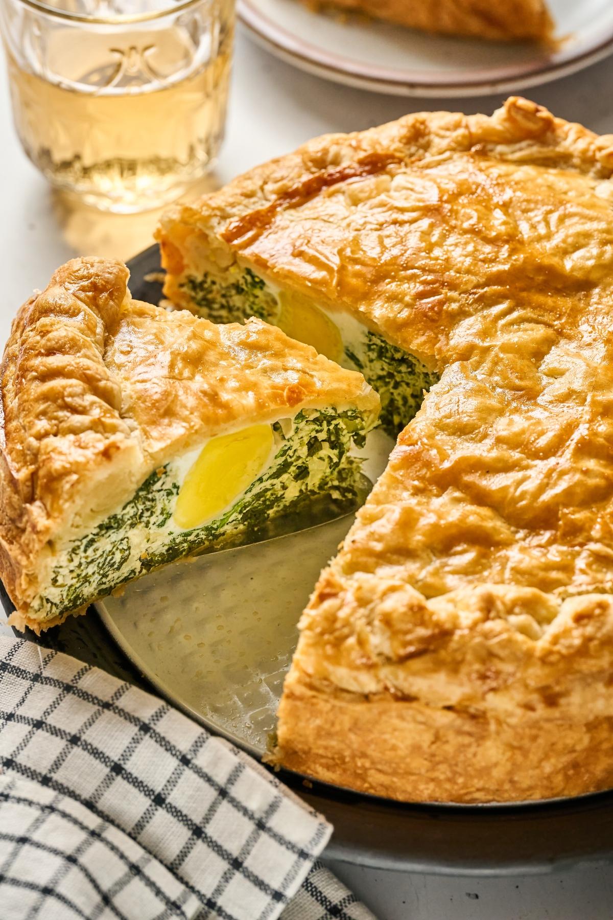 Golden spinach and egg pie with a slice being served, next to a glass of drink and a striped cloth.