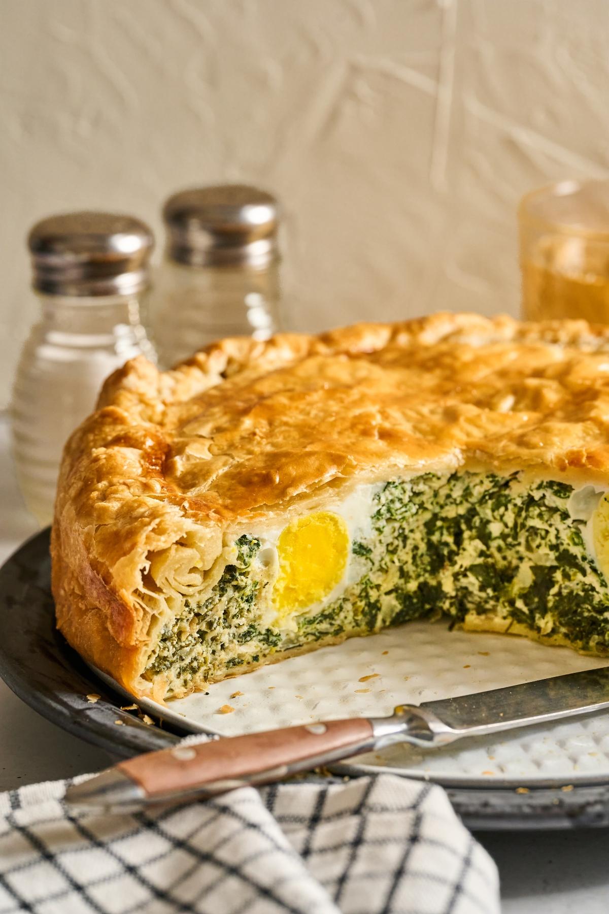 A savory pie with golden crust, filled with spinach and whole eggs, sliced open on a plate beside a knife.