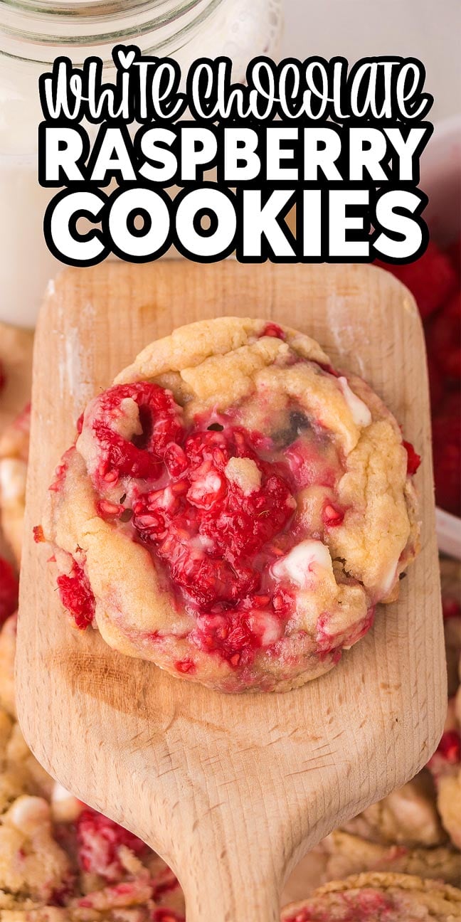 A white chocolate raspberry cookie on a wooden spatula, with more White Chocolate Raspberry Cookies and fresh raspberries in the background.