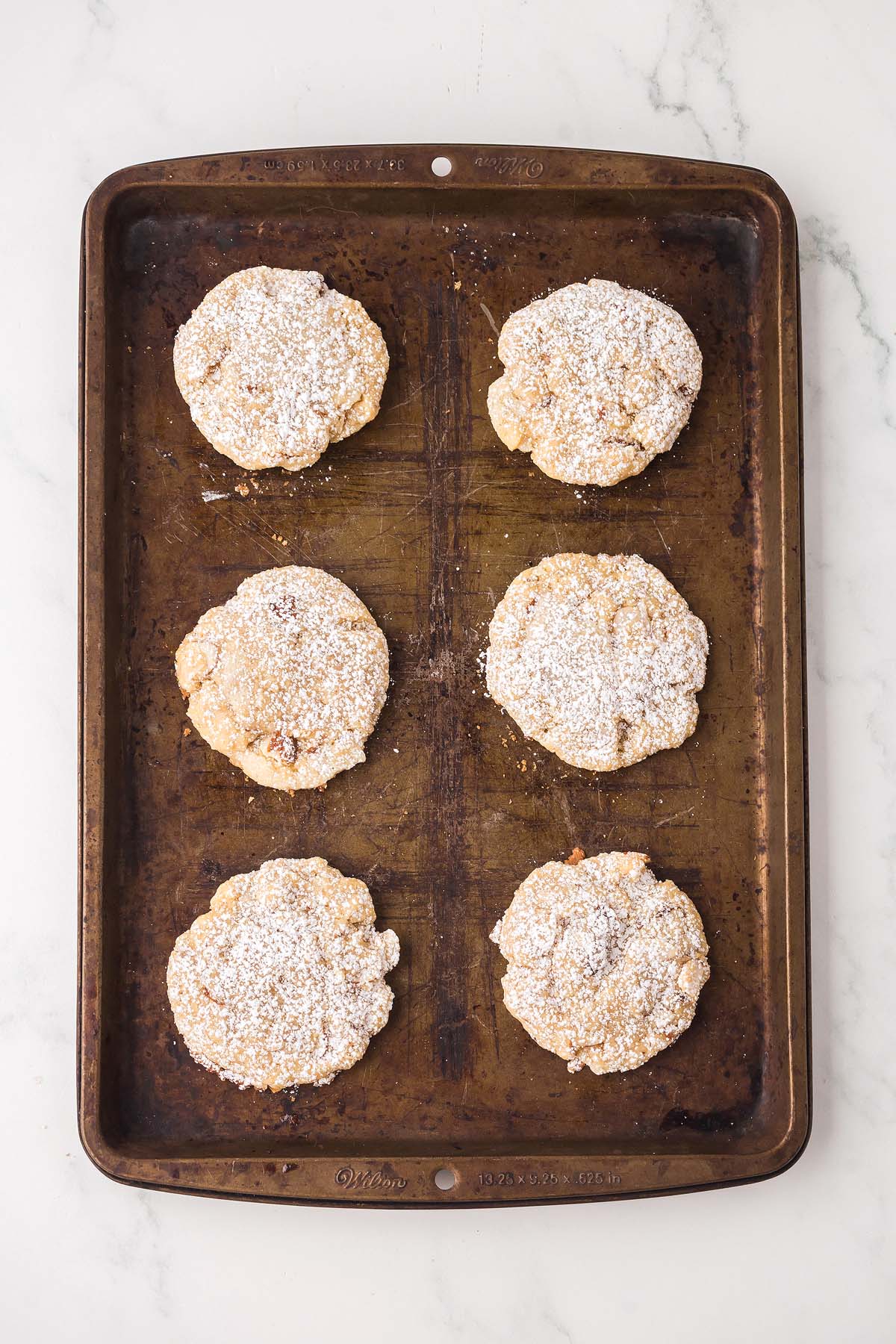 Six powdered sugar-topped Almond Croissant Cookies rest on a metal baking sheet, placed on a white marble surface.