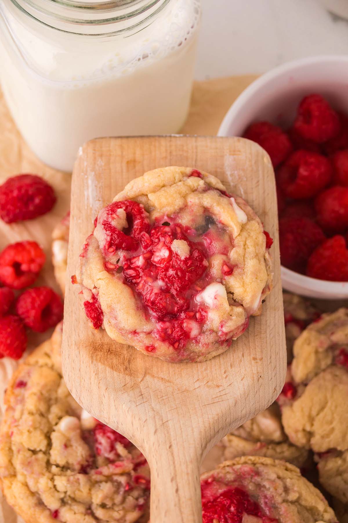 A wooden spatula holds a White Chocolate Raspberry Cookie near a bowl of raspberries and a jar of milk.